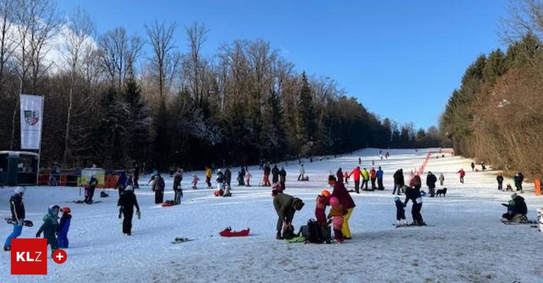 Eine Gruppe von Menschen versammelt sich auf einem verschneiten Hang, einige skifahren und andere stehen. Bäume und ein klarer blauer Himmel sind im Hintergrund.