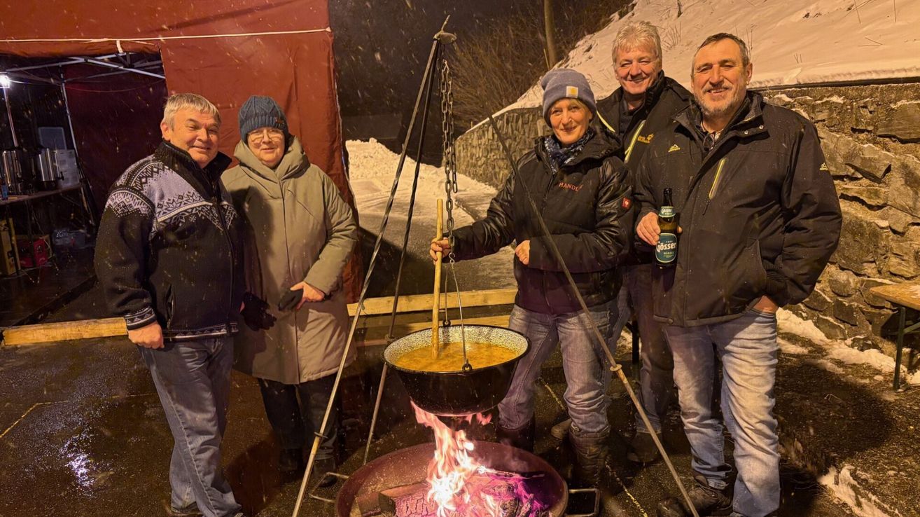 Fünf Erwachsene stehen um einen großen Topf über einem Feuer und lächeln. Einer hält einen Stock, ein anderer ein Bier. Sie sind warm gekleidet in Jacken und Mützen.