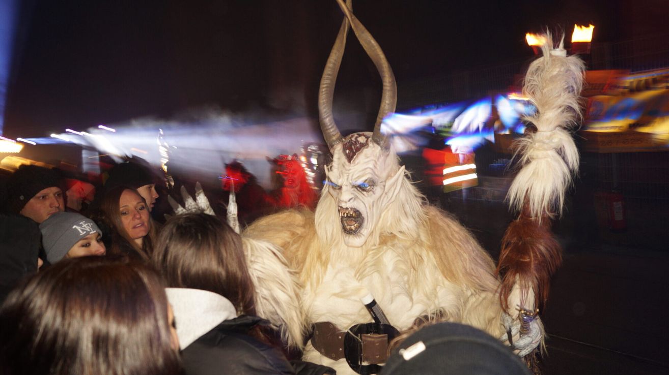 Eine Person in einem Monsterkostüm mit langen Hörnern und blauen Augen steht in der Nacht vor einer Menge.