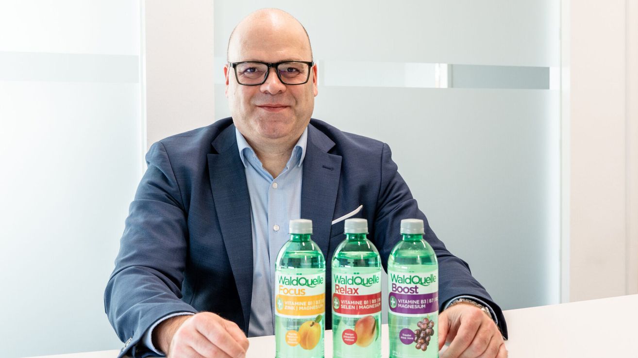 Ein Mann in einem blauen Anzug sitzt mit drei Flaschen WaldQuelle-Wasser lächelnd am Tisch.
