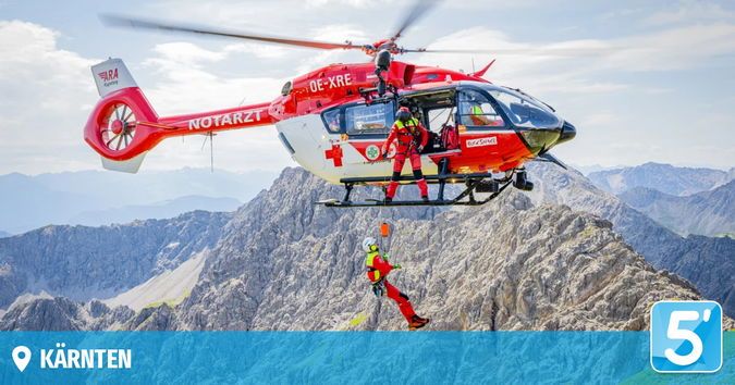 Ein roter Hubschrauber mit der Markierung 'OE-XRE' und 'NOTARZT' rettet eine Person in roter Ausrüstung. Der Hubschrauber schwebt über einer Bergkette mit bewölktem Himmel. Die gerettete Person wird durch ein Seil suspendiert.