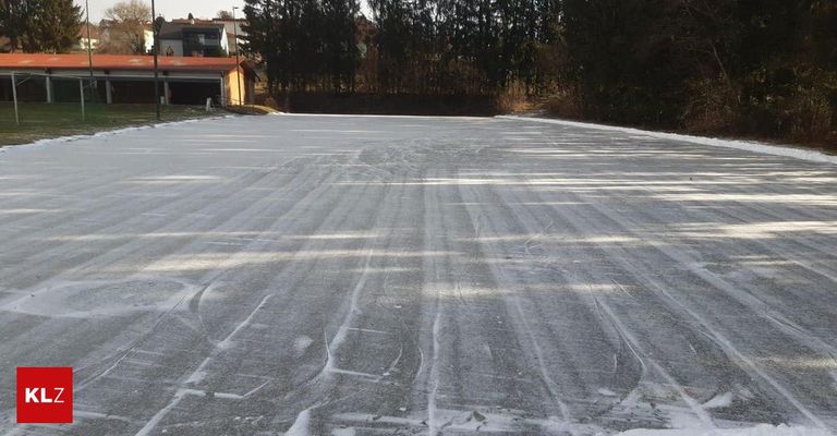 Eine gefrorene Eislaufbahn mit Reifenspuren auf dem Eis, umgeben von kahlen Bäumen und einem gelben Gebäude mit rotem Dach.