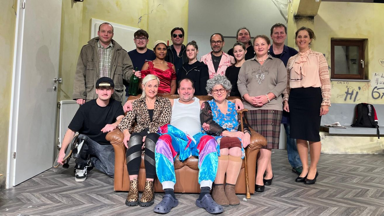 Eine Gruppe von Menschen posiert für ein Foto, einige sitzen auf einer braunen Couch, während andere dahinter stehen. Die Couch hat zwei Frauen, die darauf sitzen, eine hält einen Stock. Die Gruppe umfasst Menschen in verschiedenen Outfits und Accessoires.