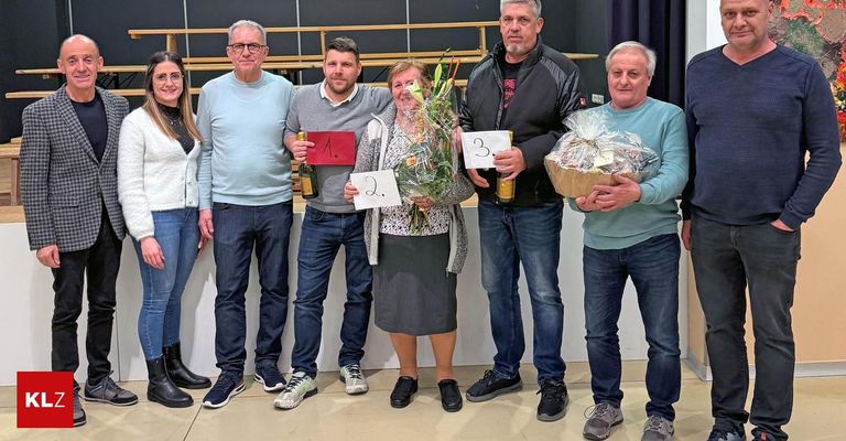Fünf Personen posieren für ein Foto mit Auszeichnungen; eine hält Blumen, eine andere eine Flasche und zwei weitere einen Korb. Sie stehen auf einer Bühne mit Bänken im Hintergrund.