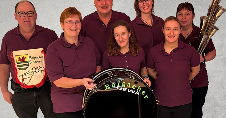 Eine Gruppe von Menschen in passenden lila Poloshirts posiert mit einem Schlagzeug, das Balgacher Gewa beschriftet ist. Die Frau vorne hält den Griff des Schlagzeugs.