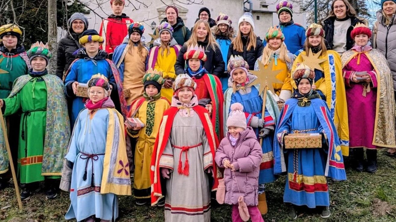 Eine Gruppe von Kindern und Erwachsenen in festlichen Kostümen posiert für ein Foto vor einem Gebäude. Einige tragen Hüte und halten Requisiten wie Sterne und Geschenke.