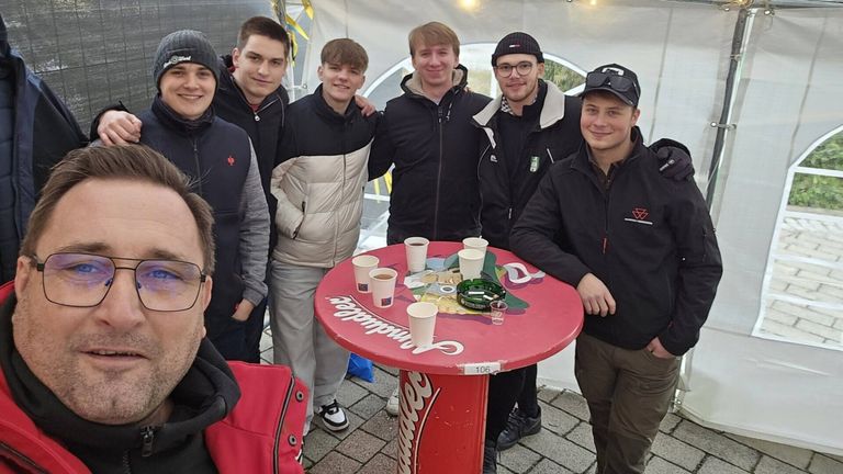 Sieben junge Männer stehen um einen runden roten Tisch mit Tassen, von denen einer einen Feuerzeug hält. Sie tragen alle Winterjacken.