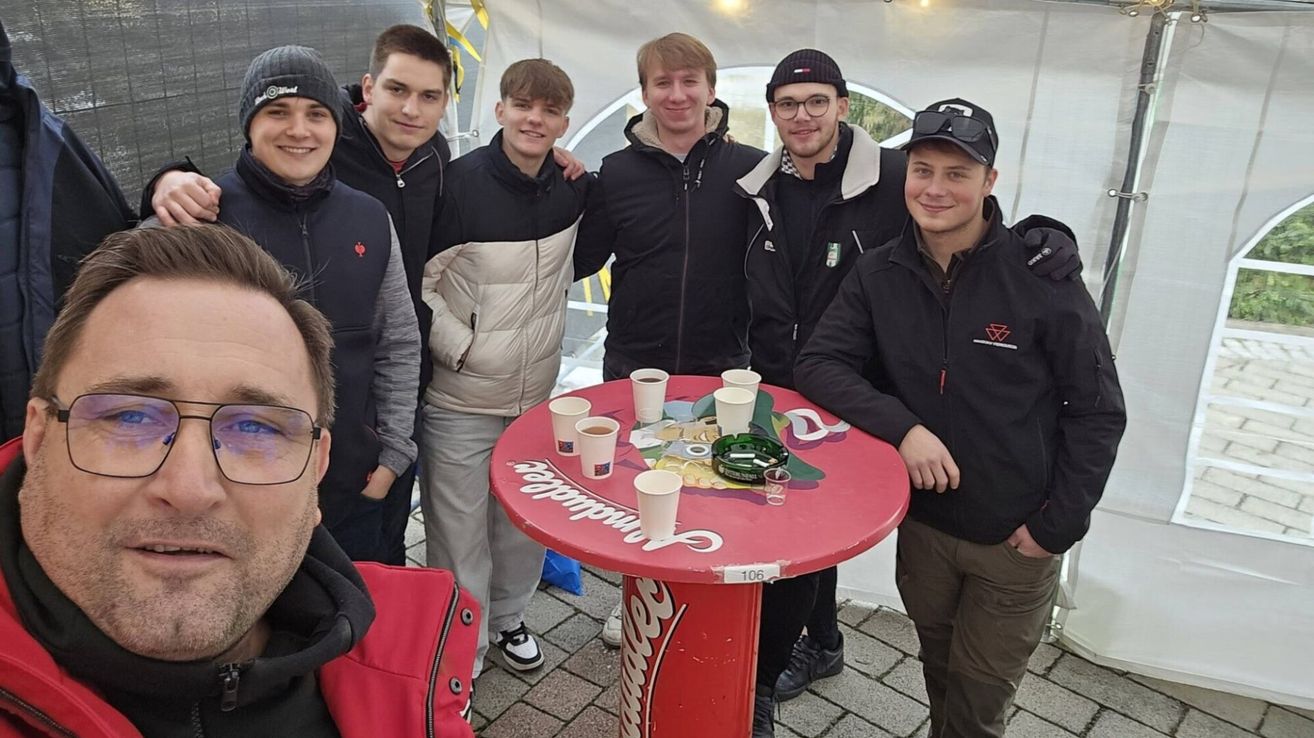 Sieben junge Männer stehen um einen runden roten Tisch mit Tassen, von denen einer einen Feuerzeug hält. Sie tragen alle Winterjacken.