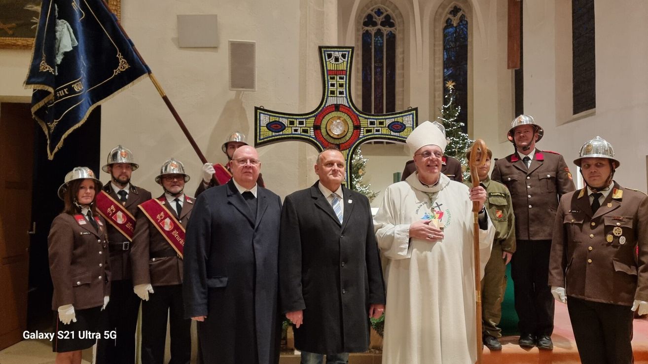Drei Männer in formeller Kleidung stehen vor einem Kirchenkreuz, ein Priester in weißen Roben und einem Bischofsstab im Vordergrund. Hinter ihnen stehen zwei Männer in Militäruniformen, einer hält eine zeremonielle Lanze. Im Hintergrund ist ein geschmückter Weihnachtsbaum zu sehen.