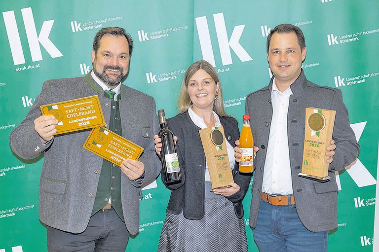 Drei Personen posieren für ein Foto und halten Auszeichnungen und Flaschen. Sie stehen vor einem grünen Hintergrund mit weißem Text. Der Mann links hält zwei Auszeichnungen und eine Flasche. Die Frau in der Mitte hält eine Trophäe und eine Flasche. Der Mann rechts hält eine Auszeichnung und eine Flasche.