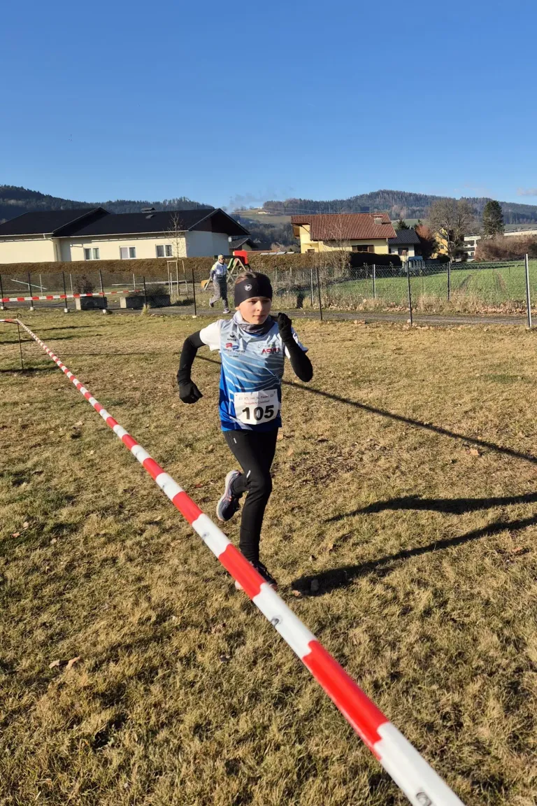 Ein junger Läufer in einem Marathon mit Kopfband, Handschuhen und einem Trikot mit der Nummer 105, der auf einem Grasfeld läuft. Im Hintergrund befinden sich Häuser und ein Zaun.