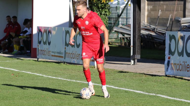 Ein Fußballspieler in einem roten Trikot dribbelt den Ball auf dem Spielfeld. Das Trikot des Spielers hat die Aufschrift 'HELD & FRANCK'.