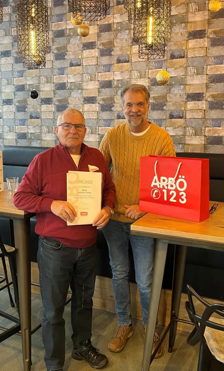 Zwei Männer stehen in einem Restaurant, einer hält ein Zertifikat und der andere lächelt. Eine rote Tasche mit der Nummer 123 steht auf dem Tisch.