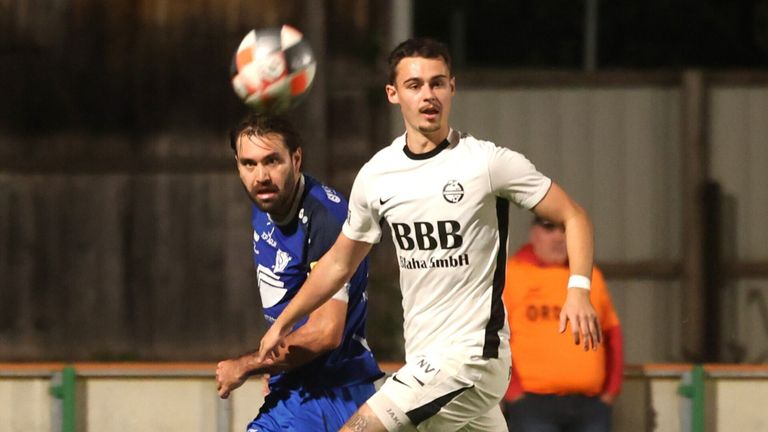 Zwei Fußballspieler konkurrieren auf einem Feld. Ein Spieler trägt ein weißes Trikot und der andere ein blaues. Sie versuchen beide, den Ball zu kontrollieren, der in der Luft ist.