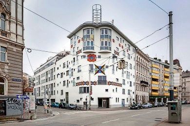 Eine urbane Straßenszene zeigt ein großes, weißes Mehrfamilienhaus mit einem dekorativen Muster an seiner Fassade. Es hat viele Fenster und steht an der Ecke einer belebten Straße mit geparkten Autos und überhängenden Straßenbahndrähten.