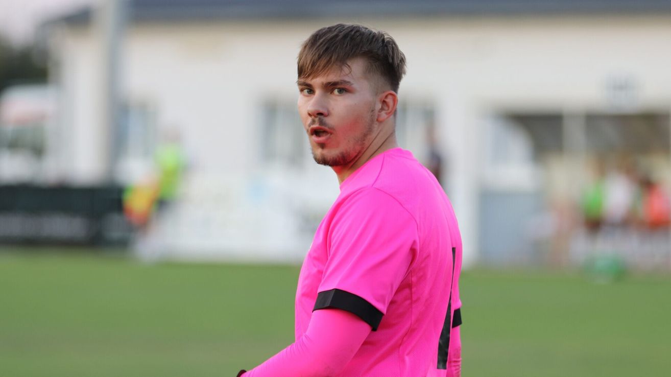 Ein Mann in einem rosafarbenen Trikot mit schwarzen Akzenten steht auf einem Feld und schreit. Dahinter ist ein verschwommenes Bild eines Gebäudes zu sehen.