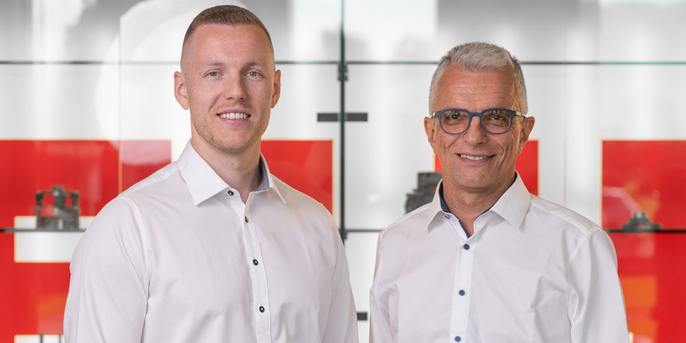 Zwei Männer in weißen Hemden mit Knöpfen lächeln vor einer Glaswand mit roten Quadraten.