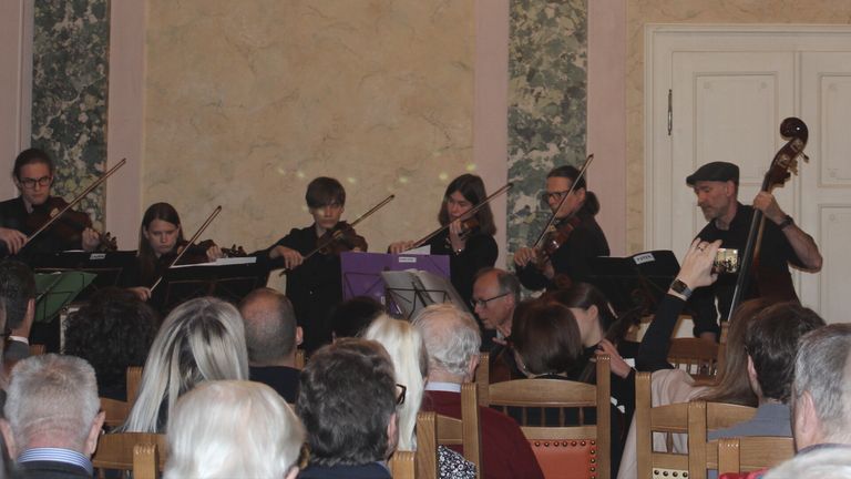 Ein Streichquartett spielt in einem schwach beleuchteten Raum, mit drei Geigern und einem Cellisten. Sie werden von einem Pianisten begleitet. Das Publikum sitzt vor ihnen.