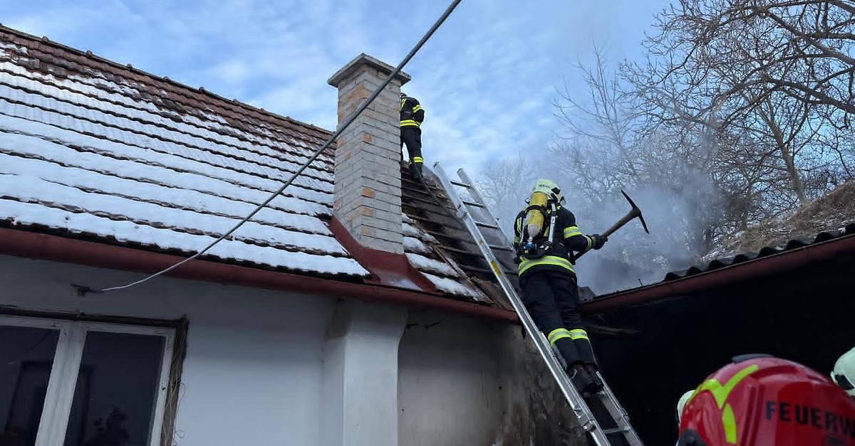 Zwei Feuerwehrleute sind auf einem schneebedeckten Dach, einer klettert eine Leiter hinauf und der andere hält eine Axt. Sie scheinen einen Schornstein an einem Haus zu bearbeiten.