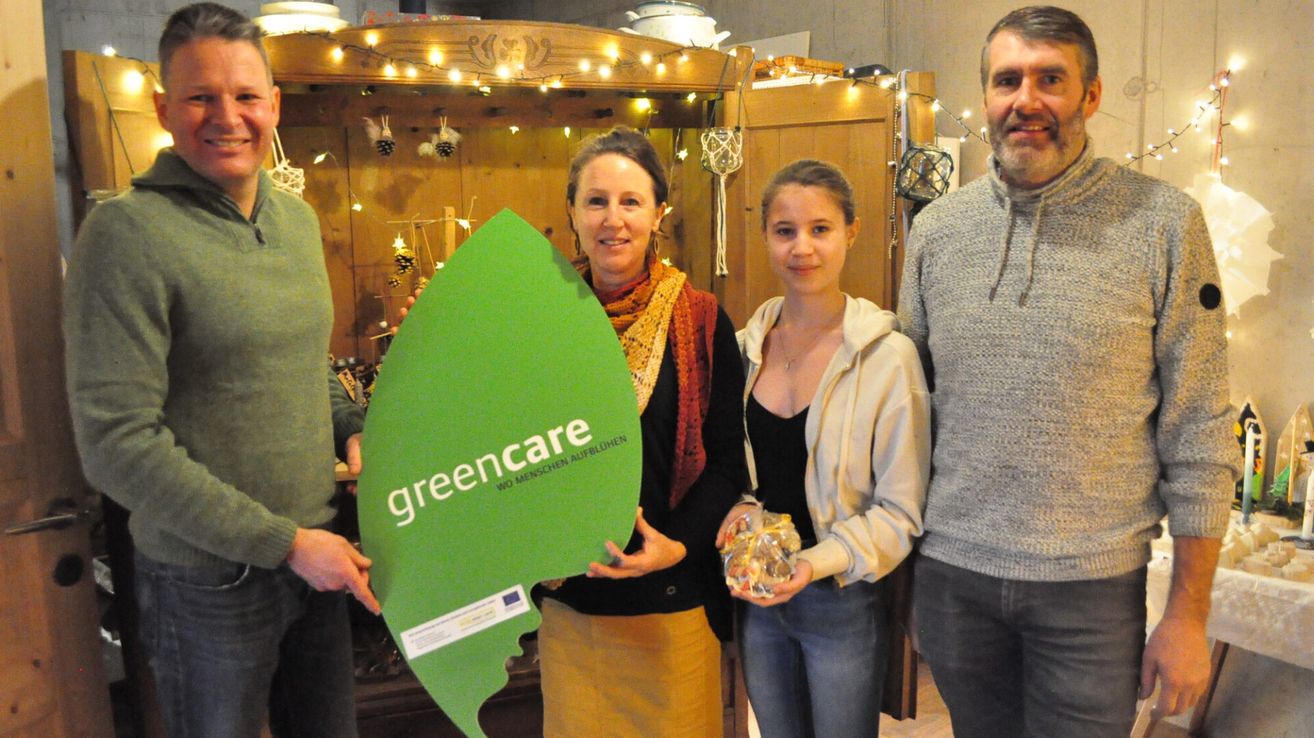 Eine Gruppe von vier Menschen steht drinnen zusammen. Eine Frau hält ein großes grünes Schild mit der Aufschrift 'greencare'. Die Frau neben ihr hält eine Tüte mit Süßigkeiten. Der Mann links und rechts hält das Schild. Hinter ihnen steht ein Holzschrank mit Lichtern und Dekorationen.