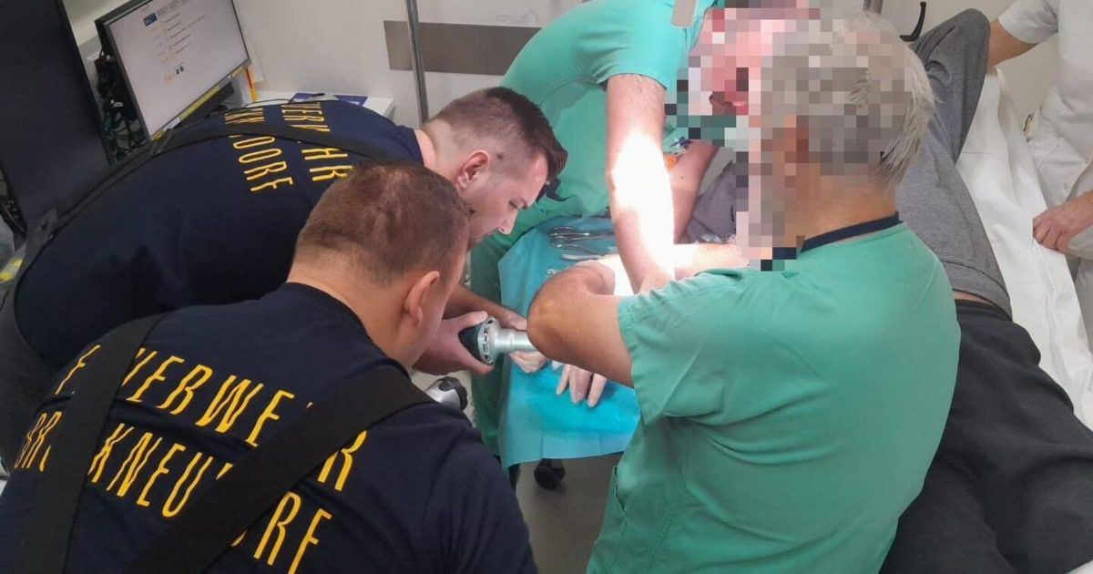 Drei medizinische Mitarbeiter in grünen Scrubs versorgen einen Patienten in einem Krankenhauszimmer. Eine Person hält ein medizinisches Werkzeug, und eine andere hat ein verschwommenes Gesicht.