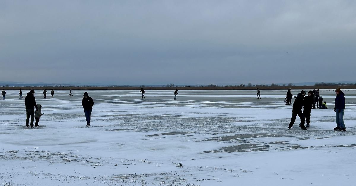 Mehrere Menschen gehen auf einem zugefrorenen See unter einem bewölkten Himmel. Sie sind in Winterkleidung gekleidet.