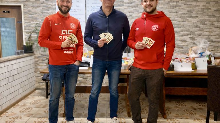 Drei Männer in Sportbekleidung, zwei in Rot und einer in Blau, stehen zusammen und halten Spielkarten, lächeln, mit einer gefliesten Wand und einem Tisch dahinter.
