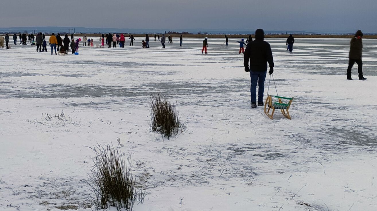 Eine verschneite Landschaft zeigt eine Gruppe von Menschen, einige auf Schlitten, mit einer Person in einer schwarzen Jacke, die einen Schlitten mit grünem Sitz zieht.