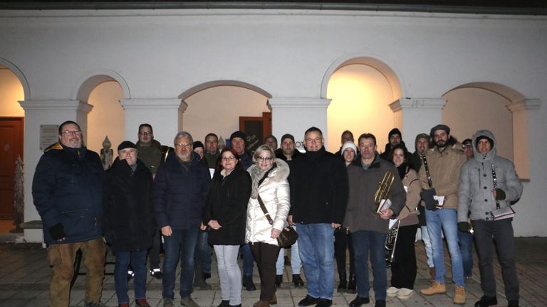 Eine Gruppe von Menschen steht draußen in Winterkleidung, einige mit Musikinstrumenten. Sie lächeln und scheinen für ein Foto zu posieren. Der Hintergrund besteht aus einem gewölbten Eingang und weißen Wänden.