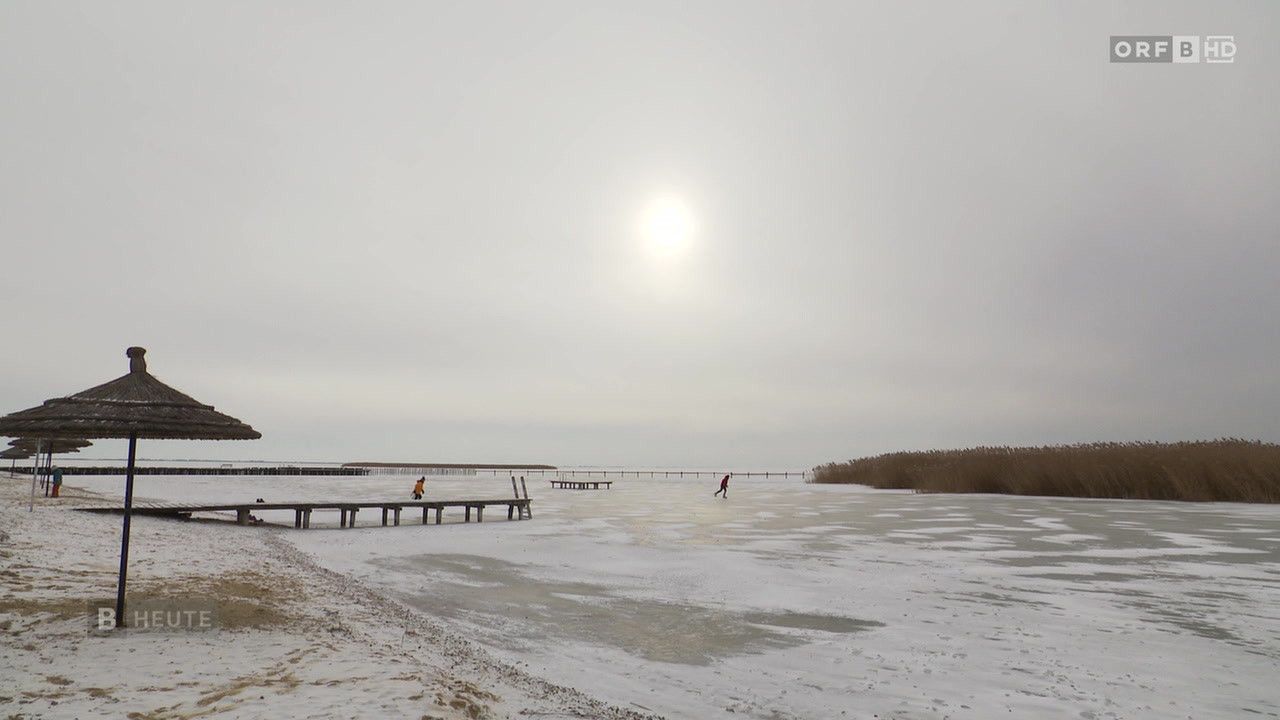 Ein sonniger Tag mit zwei Personen, die auf einem zugefrorenen See gehen, in der Nähe eines Holzstegs und Schilfrohrs.