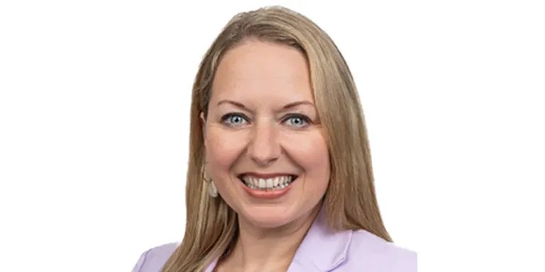 Eine Frau mit blonden Haaren, blauen Augen und einem lächelnden Gesicht trägt ein lila Hemd. Sie schaut fröhlich nach vorne.