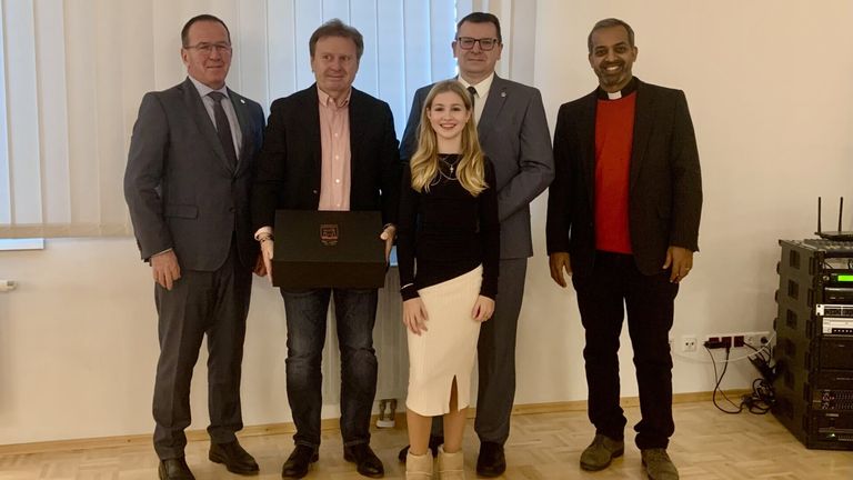 Fünf Männer und eine Frau stehen zusammen für ein Foto. Die Frau hält eine Box und trägt eine Halskette. Ein Mann trägt ein rotes Hemd. Sie sind in einem Raum mit einer weißen Wand und einem Holzboden.