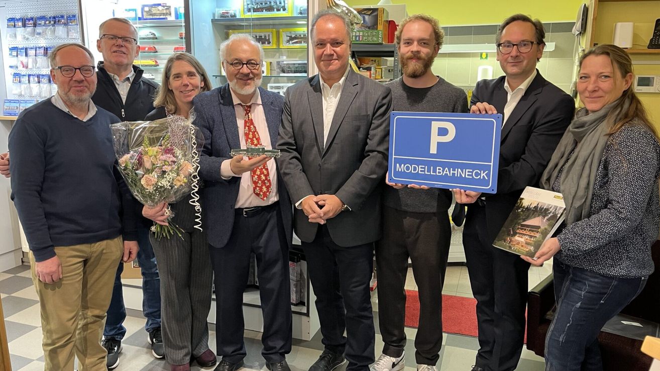 Fünf Personen stehen in einem Raum mit einem Ladenhintergrund. Sie halten ein Schild mit 'P' und 'ModellBahneck'. Der Mann auf der linken Seite hält einen Blumenstrauß.