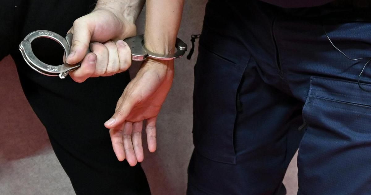 Eine Person in Handschellen wird von einer anderen Person, wahrscheinlich einem Beamten, festgehalten, wobei eine Hand am Arm und die andere die Handschellen hält.