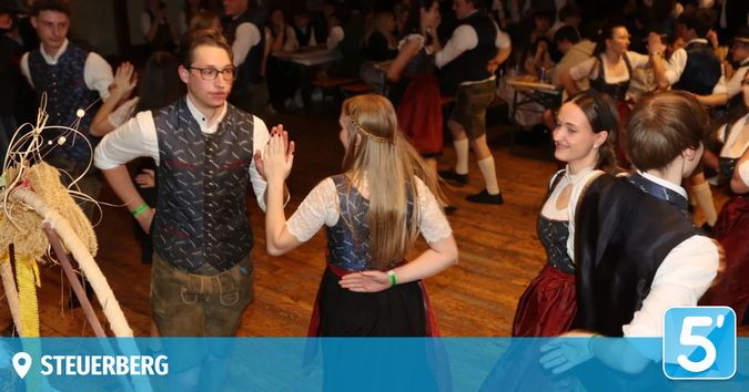Mehrere Menschen tanzen auf einem Holzboden in traditionellen Kostümen, ein Mann mit Brille und die Frauen mit langen blonden Haaren und einer Blume im Haar.
