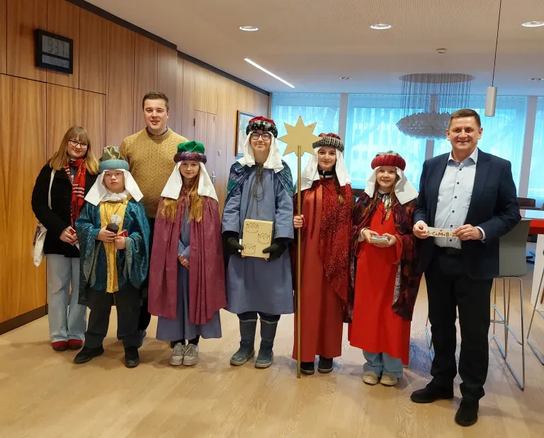 Eine Gruppe von Menschen in mittelalterlichen Kostümen posiert für ein Foto. Die Kostüme bestehen aus Kronen, Schals und Roben. Eine Person hält einen goldenen Stern und ein Buch. Das Foto wurde in einem Raum mit Holzwänden und einer Kristallleuchter aufgenommen.