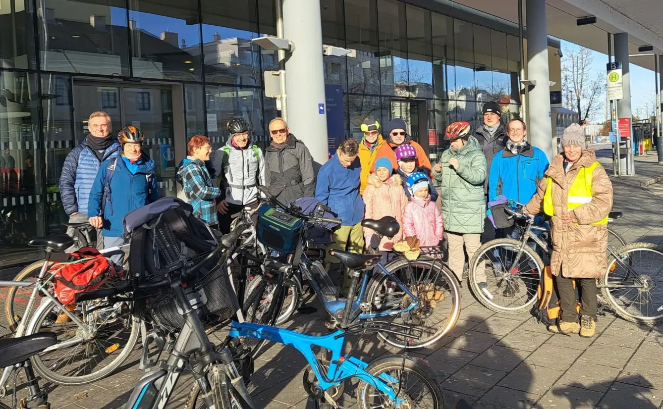 Eine Gruppe von Radfahrern, einige mit Helmen, steht vor einem modernen Gebäude. In der Nähe sind Fahrräder in verschiedenen Farben geparkt, und die Radfahrer scheinen in ein Gespräch vertieft zu sein.