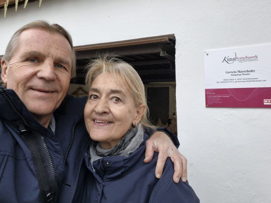 Ein älterer Mann und eine ältere Frau posieren für ein Selfie, lächelnd und stehend in der Nähe einer Wand mit einem roten Schild, das 'Kunsthaus Gerwin Mayer' liest.