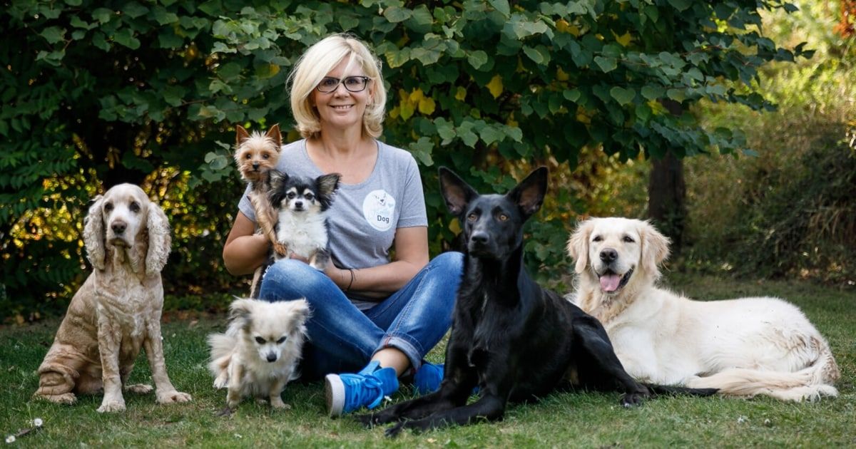 Eine Frau mit Brille und einem grauen T-Shirt mit Hundelogo sitzt mit ihren Hunden auf Gras.