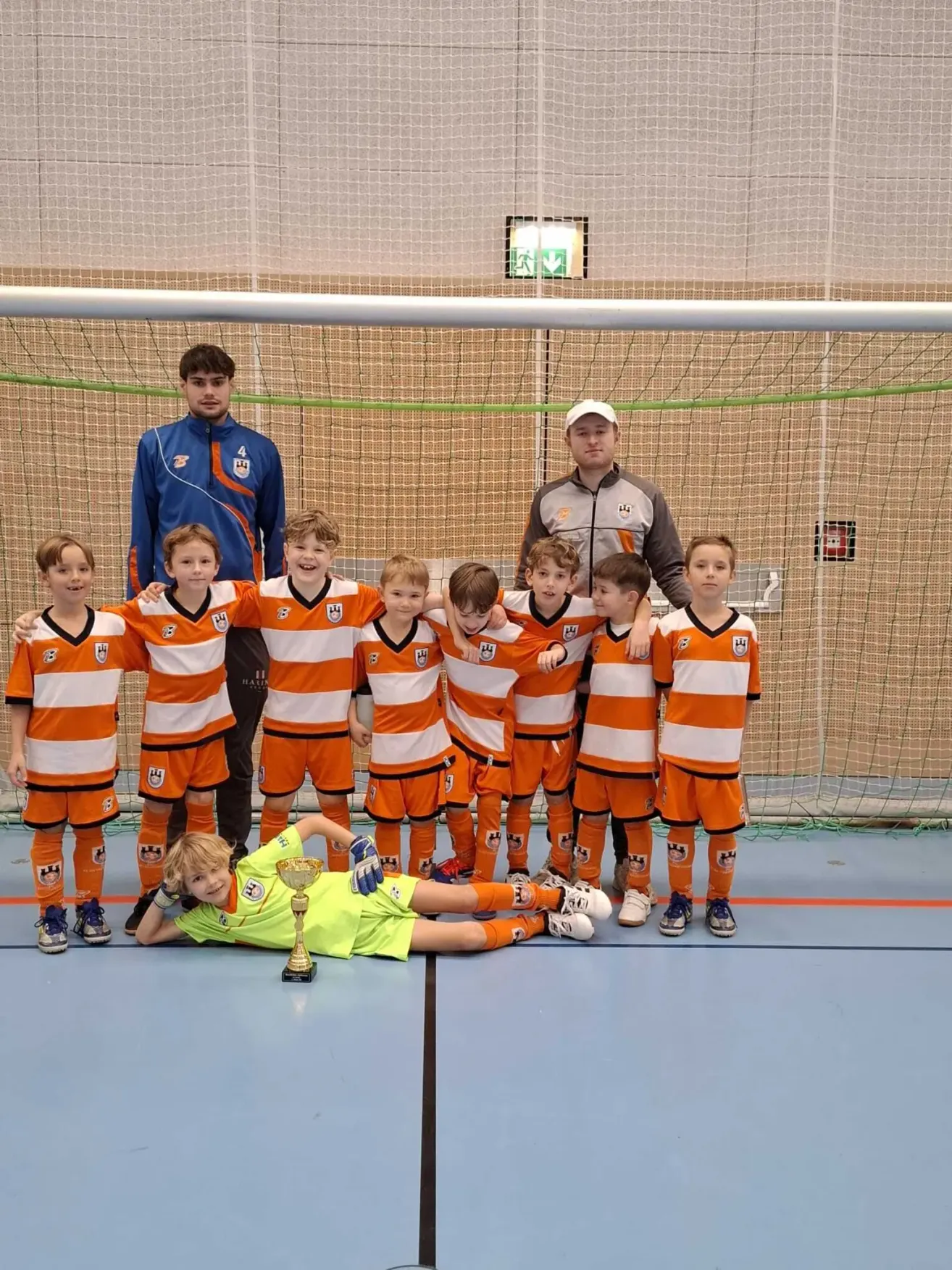 Ein Fußballteam posiert für ein Foto mit ihrem Trainer und einer Trophäe auf dem Boden. Das Team trägt orange und weiße Uniformen.