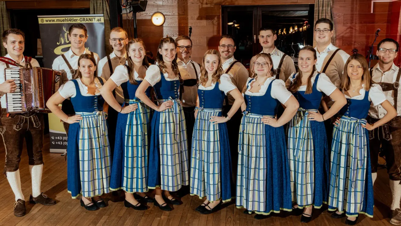Eine Gruppe von Frauen und Männern in traditioneller Kleidung posiert für ein Foto, mit Holzwänden und einer Lampe im Hintergrund.