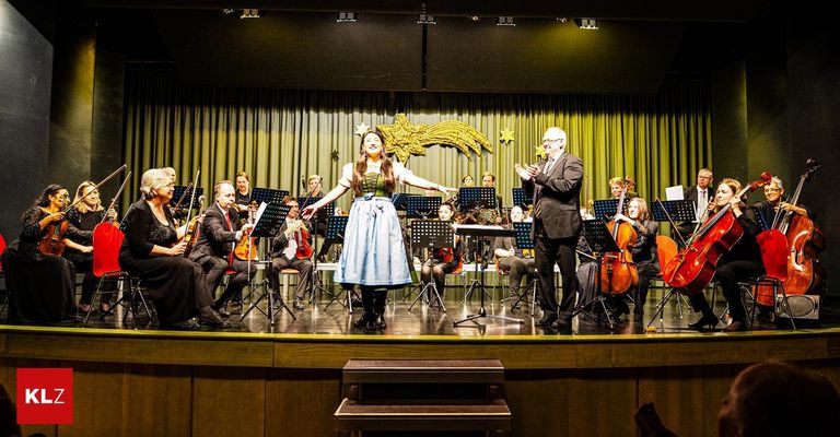 Eine Frau in traditioneller Kleidung steht auf der Bühne und gestikuliert, während ein Orchester hinter ihr spielt. Die Bühne ist mit einem Sternenhintergrund geschmückt.