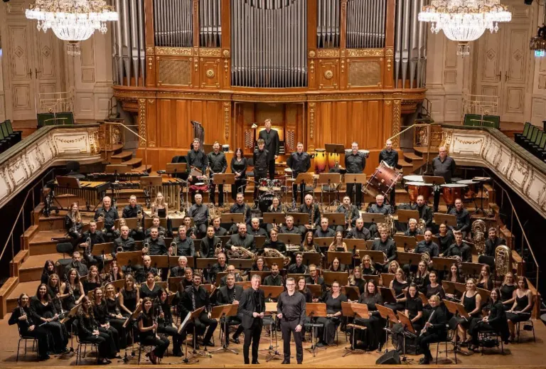 Ein großes Orchester spielt auf einer Bühne mit einer großen hölzernen Orgel im Hintergrund. Die Musiker tragen schwarze Kleidung und spielen verschiedene Instrumente. Zwei Männer stehen vor dem Orchester und dirigieren möglicherweise.