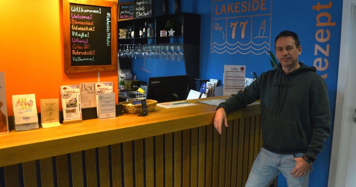 Ein Mann steht hinter einem Tresen mit einem Schild, das Lakeside 77 anzeigt. Der Tresen hat eine Tafel mit verschiedenen Willkommensbotschaften in verschiedenen Sprachen.