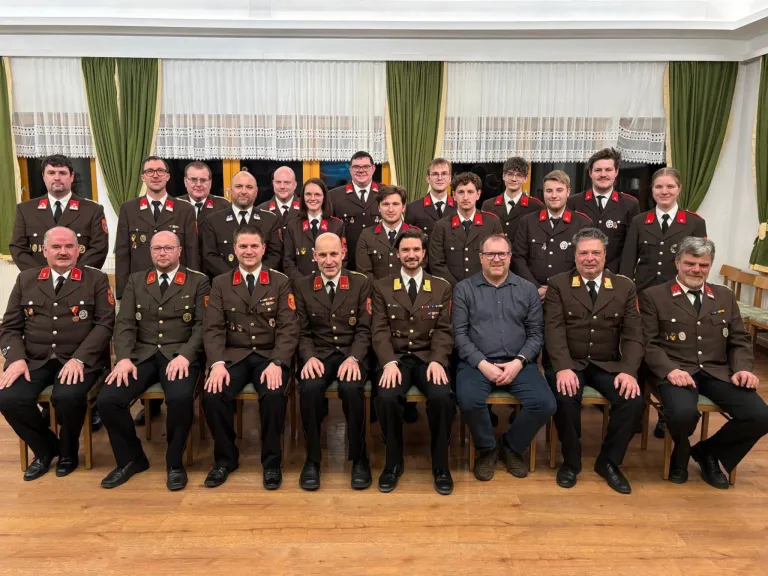 Eine Gruppe von Menschen in Militäruniformen posiert für ein Foto. Einige sitzen auf Stühlen, andere stehen. Dahinter hängen zwei grüne Vorhänge über einem Fenster.