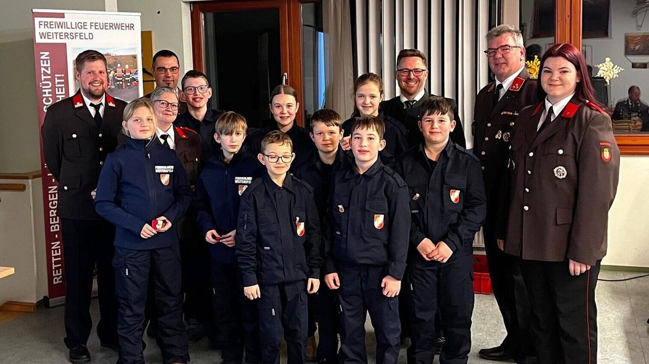 Eine Gruppe von Kindern und Erwachsenen in Uniform steht gemeinsam für ein Foto, wahrscheinlich in einer Schule oder einem Trainingsbereich. Die Kinder tragen schwarze Uniformen mit einem Logo auf der linken Brust. Die Erwachsenen sind in Militäruniformen.