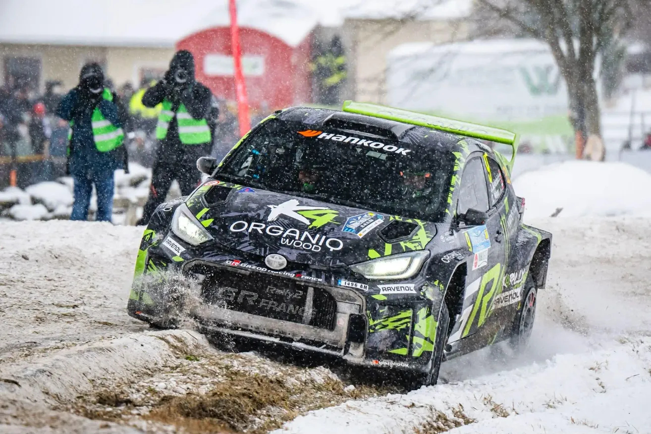 Ein Rallyeauto fährt durch den Schnee, während eine Person im Hintergrund filmt. Das Auto hat ein grünes und schwarzes Design, und das Wort Hankook steht auf dem Dach.