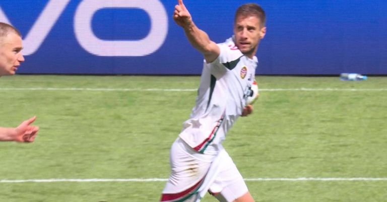 Ein Fußballspieler in weißem Trikot läuft auf dem Feld und zeigt mit seiner rechten Hand. Das Feld hat weiße Markierungen und eine blaue Wand mit einem weißen Buchstaben O.