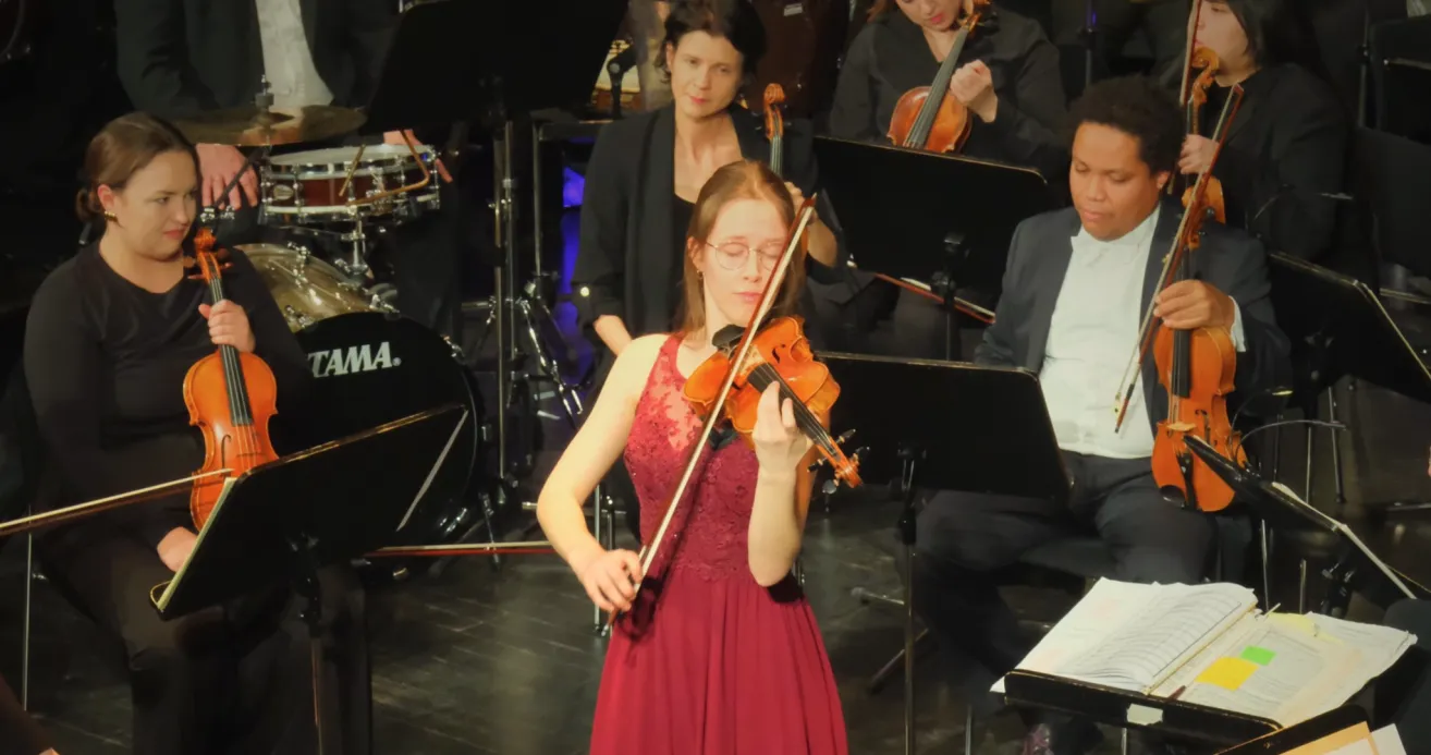 Eine Frau in einem roten Kleid spielt die Geige auf einer Bühne mit anderen Musikern. Im Hintergrund sind verschiedene Instrumente wie Cello und Schlagzeug zu sehen.