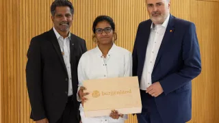 Drei Personen stehen vor einer Holzwand, zwei Männer und eine Frau. Die Frau hält eine Holzbox mit dem Wort 'Burgenland' darauf.